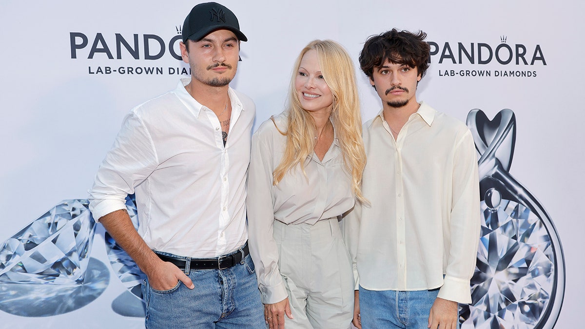 Pamela Anderson in a matching pale blue/grey blouse and trousers smiles and laughs with her two sons, wearing white shirts and jeans