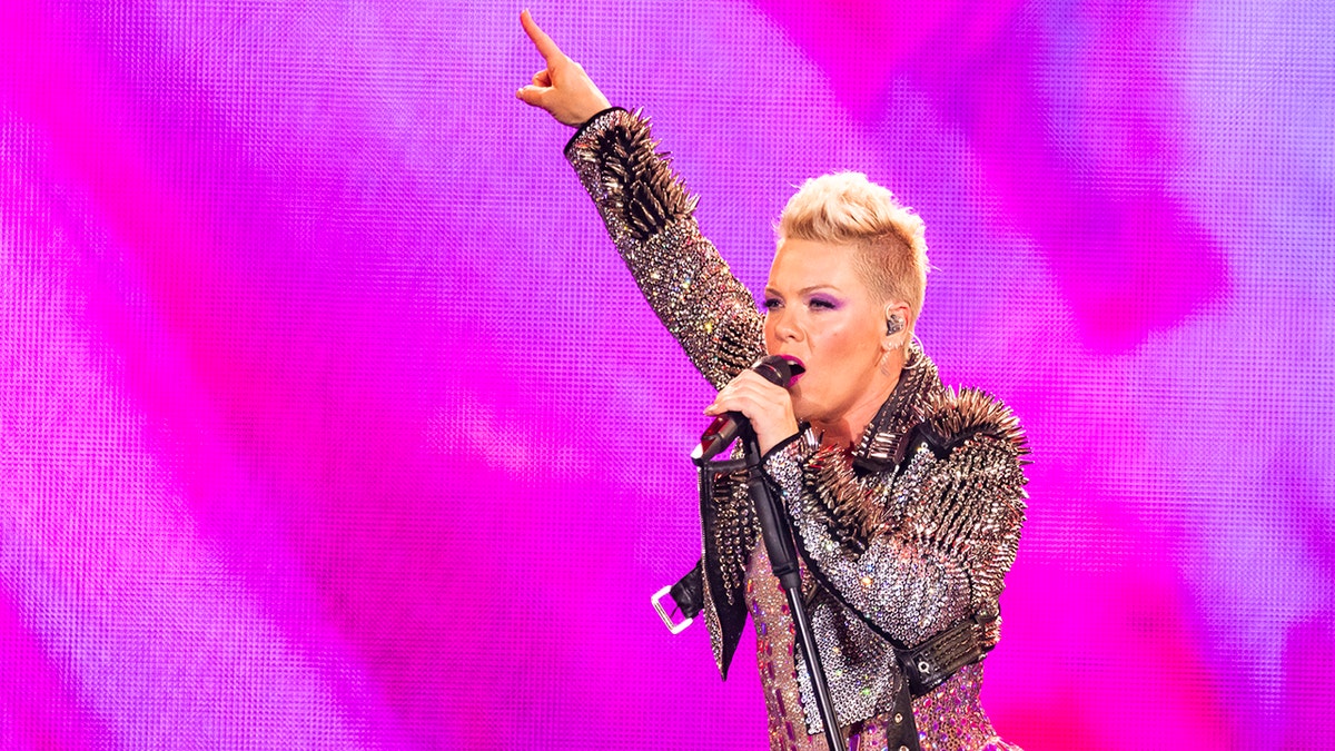 Pink aponta para o ar em frente a uma tela magenta em uma jaqueta de couro com tachas se apresentando no palco