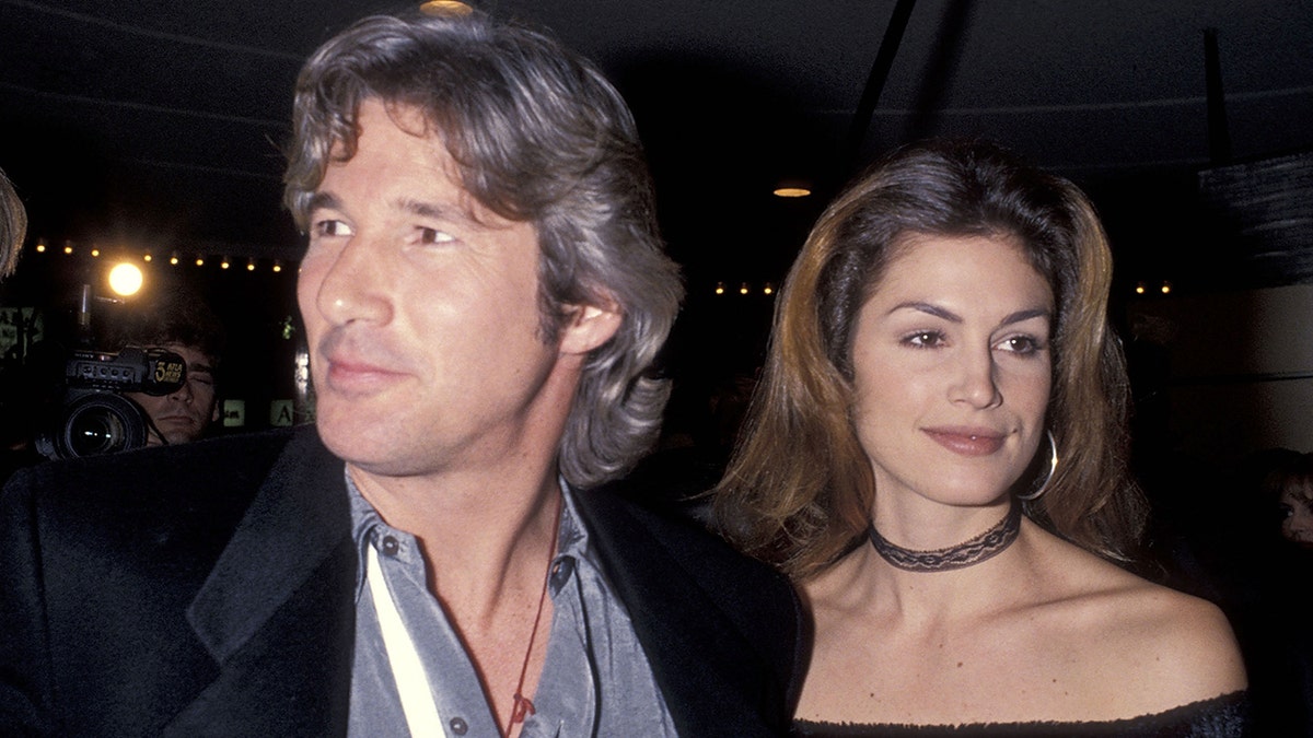 Richard Gere in a black suit looks to his right as he walks with Cindy Crawford in a black choker necklace who looks straight ahead
