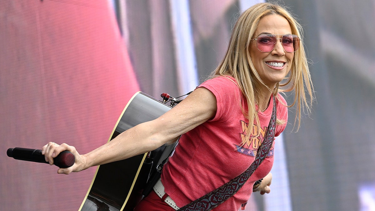 Sheryl Crow leans forward on stage in a red shirt with her guitar behind her back and red transparent sunglasses, smiling for the camera