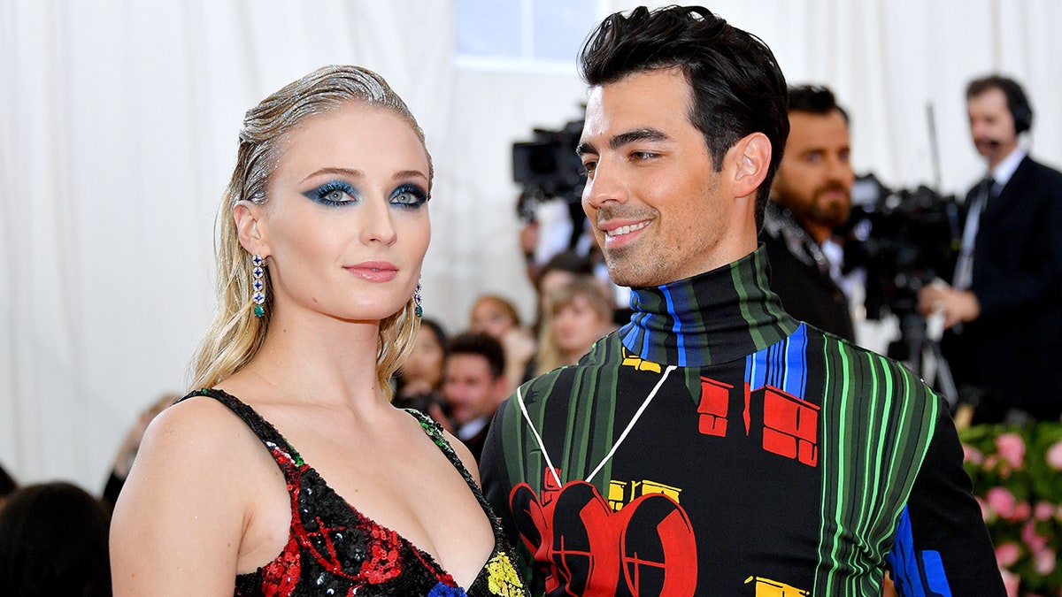 Joe Jonas in a multi-colored shirt looks longingly at Sophie Turner in a black and multi colored dress at the Met Gala in 2019