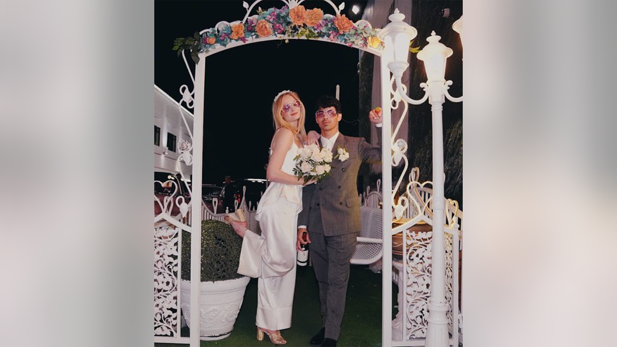 Sophie Turner popping her foot wearing a white jumpsuit under an arch with Joe Jonas at their wedding in Vegas