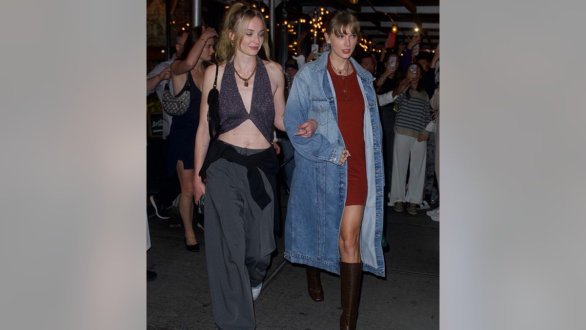 Sophie Turner em um top curto e calças de cintura baixa com um suéter amarrado nos quadris segura o braço de Taylor Swift enquanto caminhava pela cidade de Nova York