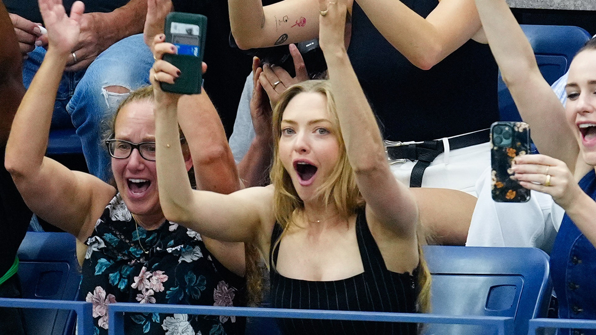 Amanda Seyfried in a black tank top raises her hands in the air, holding her phone watching the US Open