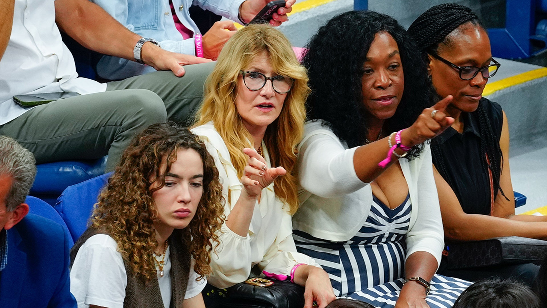 Laura Dern in a white blouse and glasses points while seated next to Shonda Rhimes in a blue striped dress and white cardigan