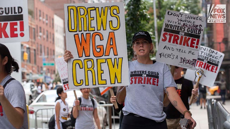 A woman holds an orange and yellow sign that reads "DREW'S WGA CREW"