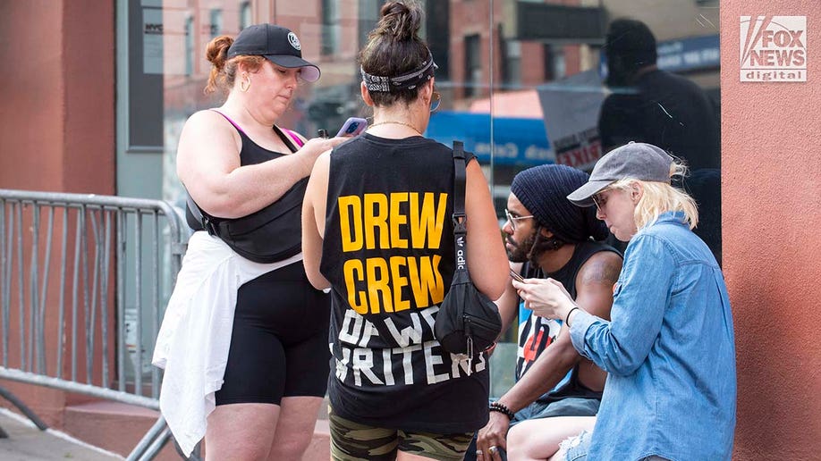 A person wears a "Drew Crew" shirt with tape added that says "OF WGA WRITERS"
