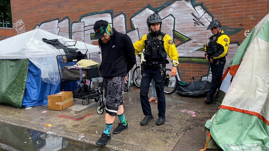 Portland bike squad officer leads a handcuffed man away from tents to a patrol vehicle