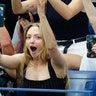 Amanda Seyfried in a black tank top raises her hands in the air, holding her phone watching the US Open