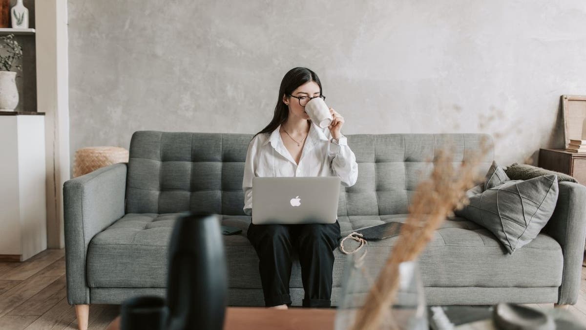 Woman sitting on a couch, sipping coffee and typing on her laptop.