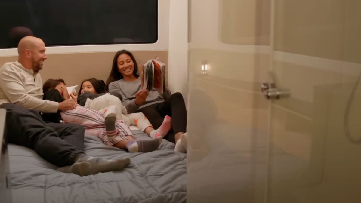 A family lays in bed together while reading a book.