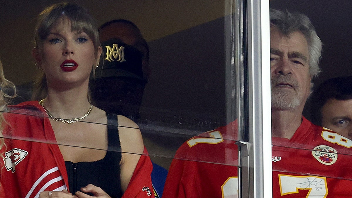 Taylor Swift in a black tank top and red jacket watches alongside Ed Kelce in a Travis Kelce Chief's jersey