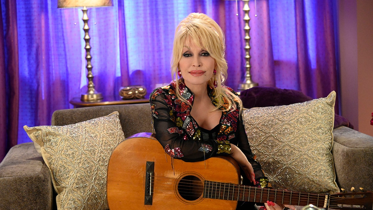 Dolly Parton sits on a couch with her guitar in her lap