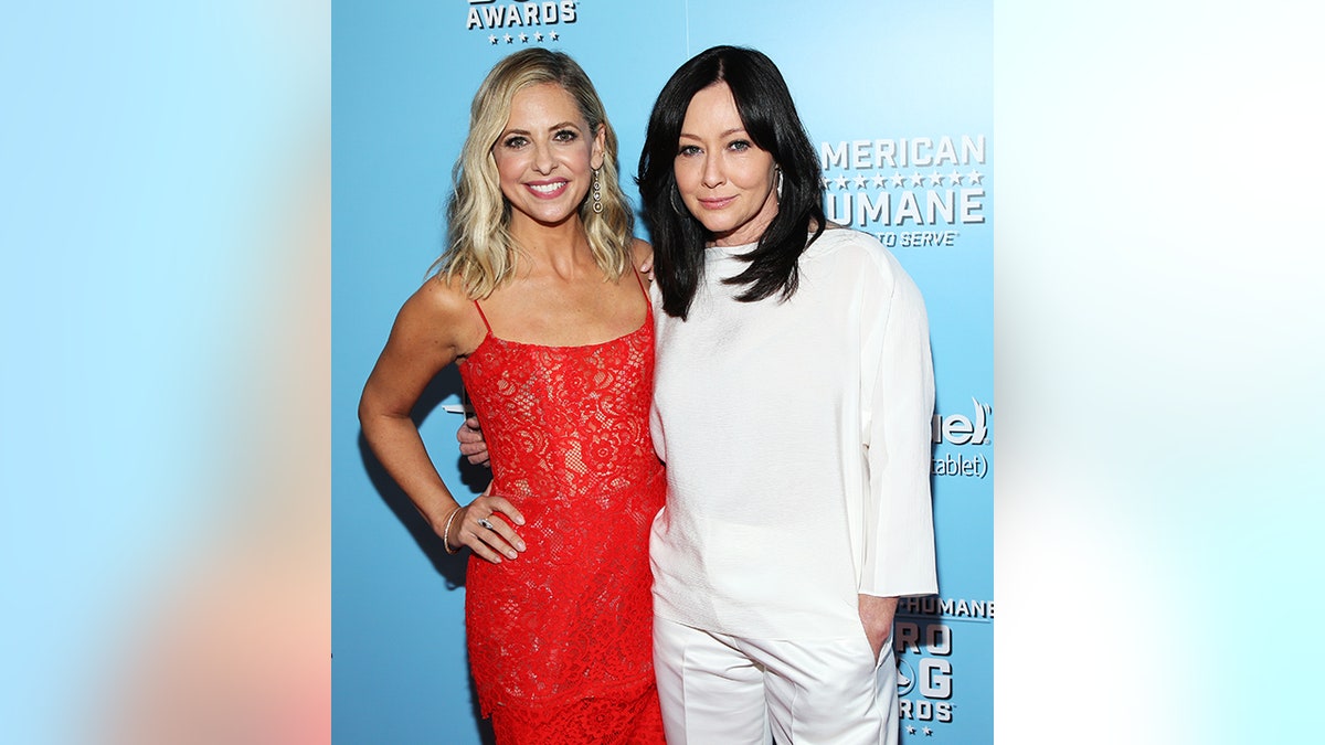 Sarah Michelle Gellar in a red dress smiles for a photo with Shannen Doherty in a white dress on the carpet
