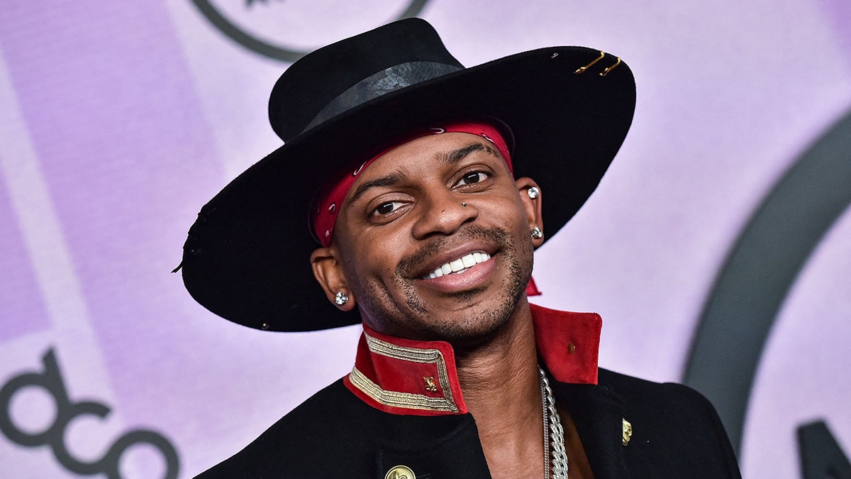 Jimmie Allen in a black hat and black jacket with red and gold trim on the American Music Awards carpet