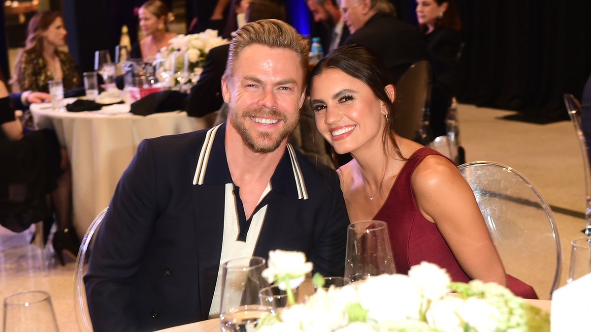 Derek Hough in dark suit smiles at a table next to Hayley Erbert in a maroon dress