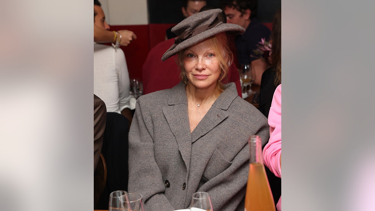 Pamela Anderson sits demurely at an event with a tilted hat