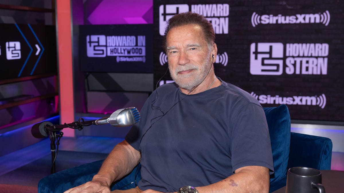 Arnold Schwarzenegger in a blue shirt sits down at a table for "The Howard Stern Show"