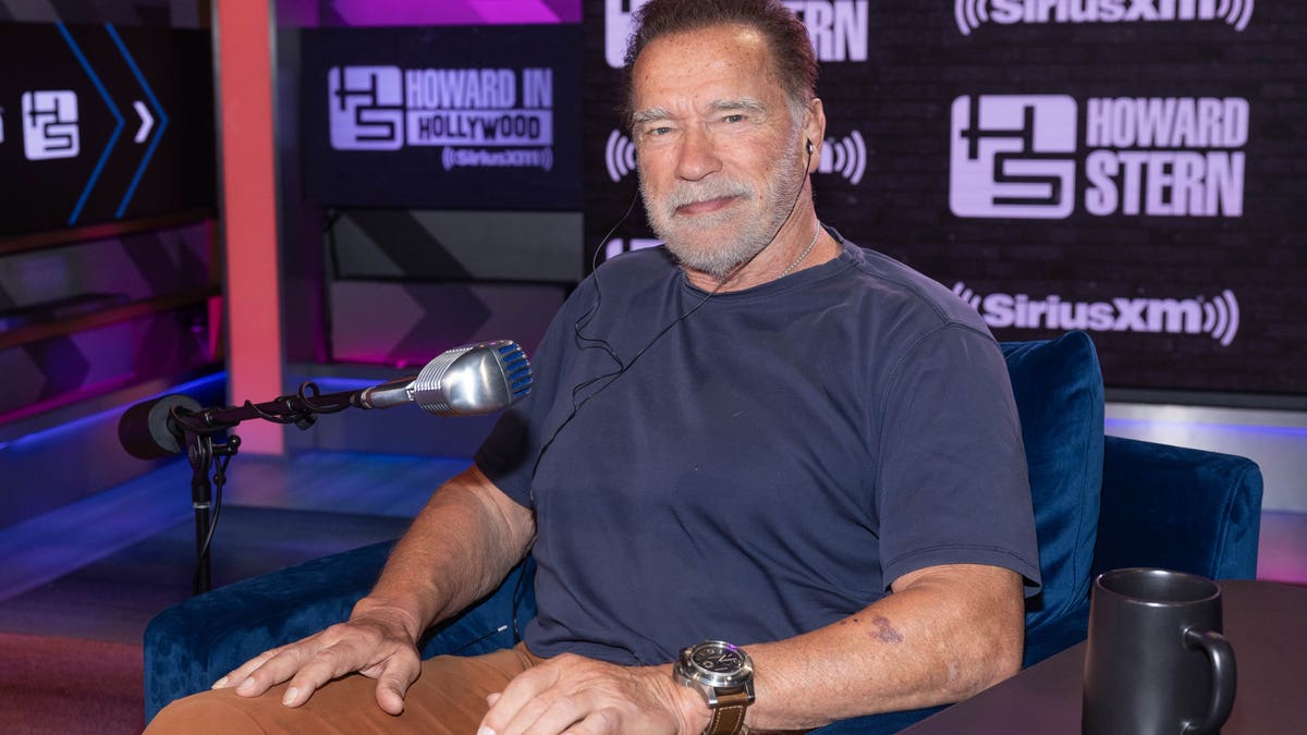 Arnold Schwarzenegger in a blue shirt sits down at a table for "The Howard Stern Show"