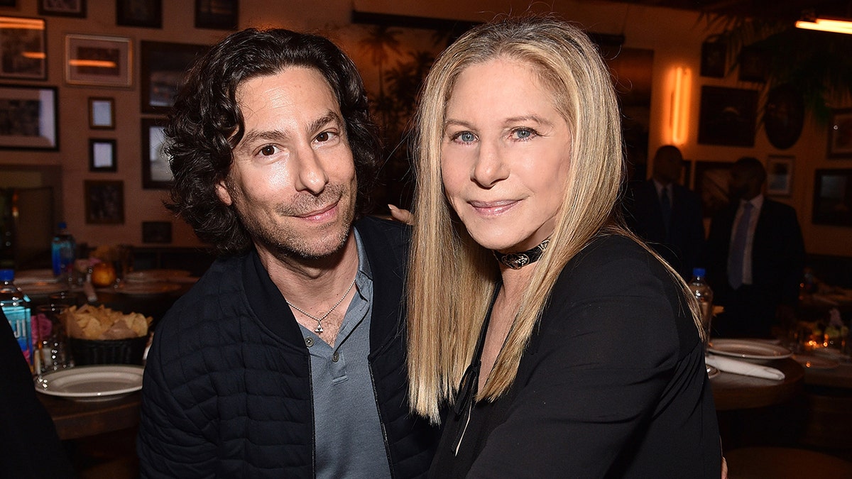 Jason Gould and Barbra Streisand leaning against each other and smiling as they wear matching black outfits