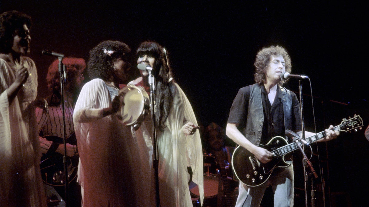 Bob Dylan playing on a guitar as a group of female gospel singers stand next to him on stage