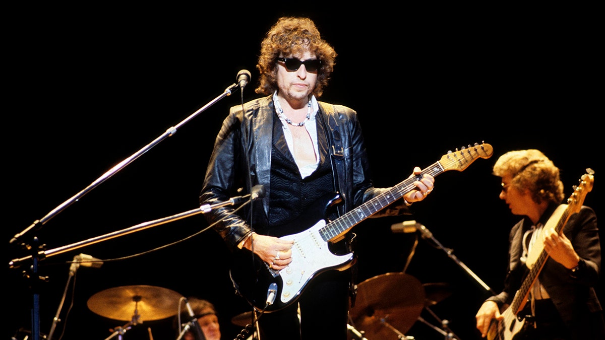 Bob Dylan holding an electric guitar and looking serious with sunglasses on stage