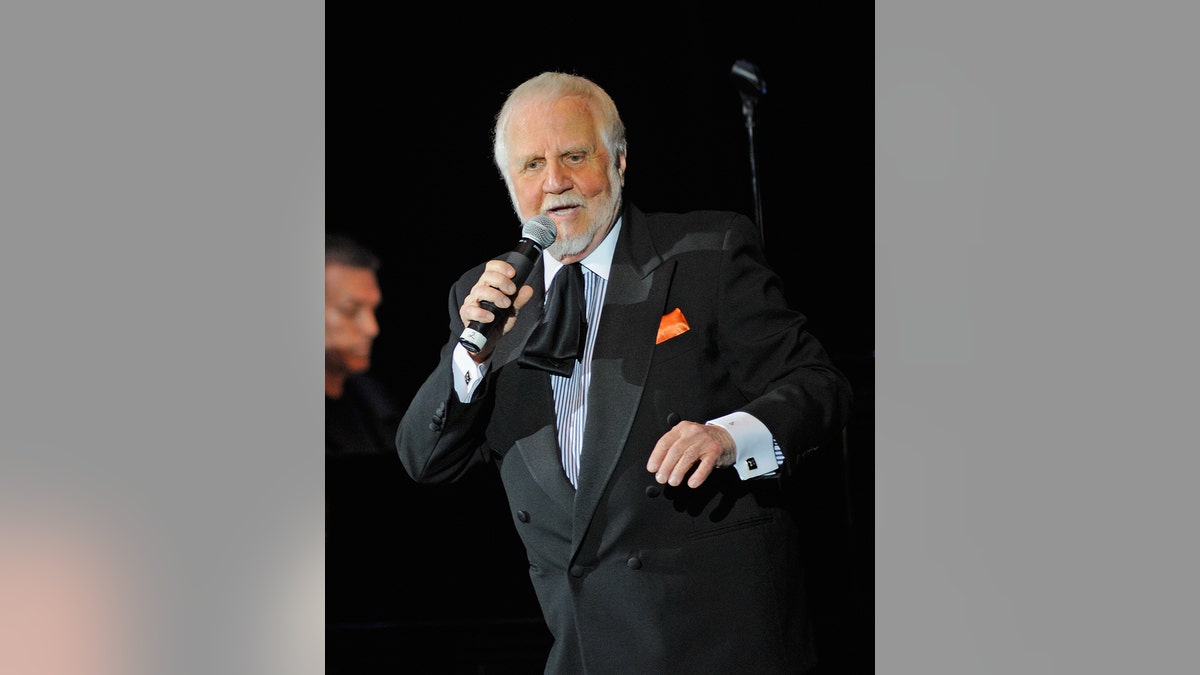 Bruce Belland wearing a dark suit and singing to a mic
