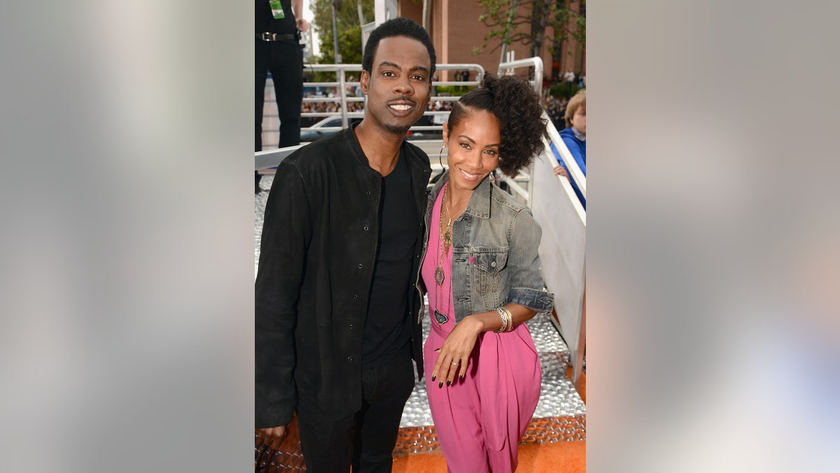 Chris Rock wearing an all black outfit and Jada Pinkett Smith wearing a pink jumpsuit and a denim jacket