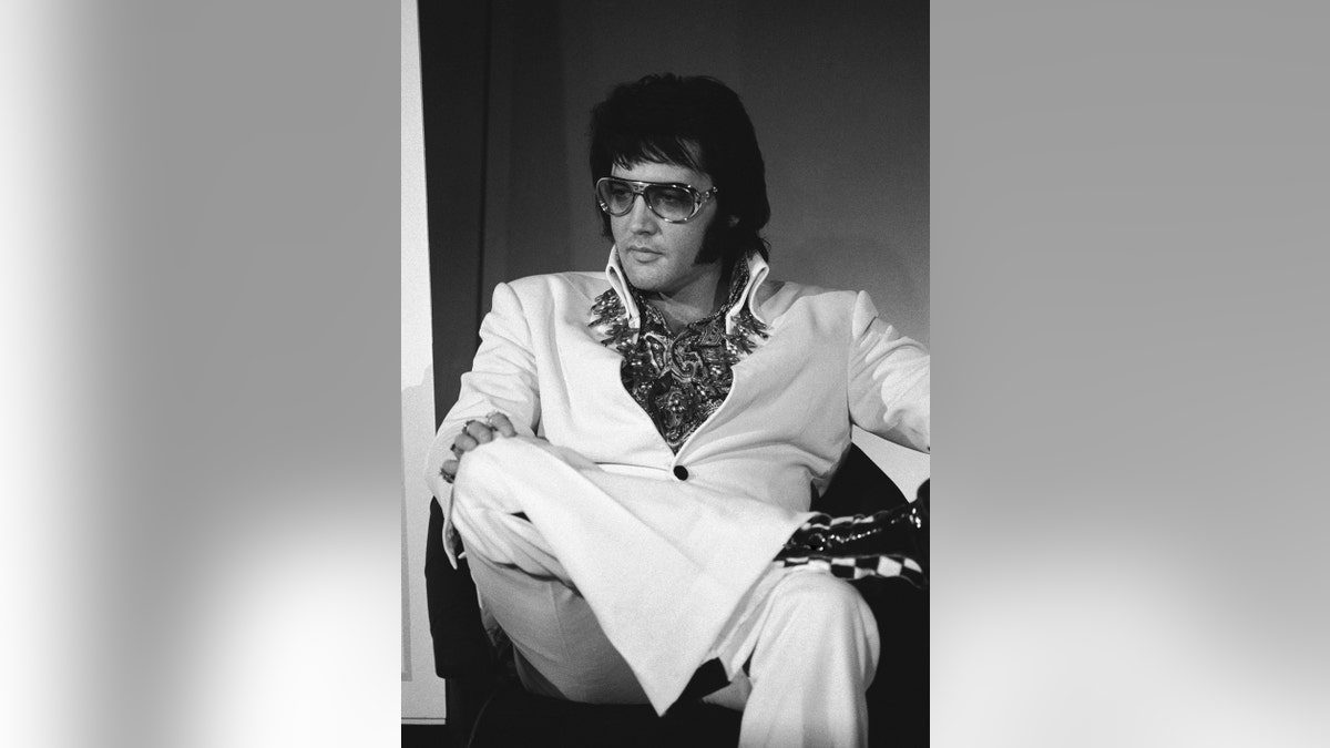 Elvis Presley sitting down and wearing a white jumpsuit with sunglasses