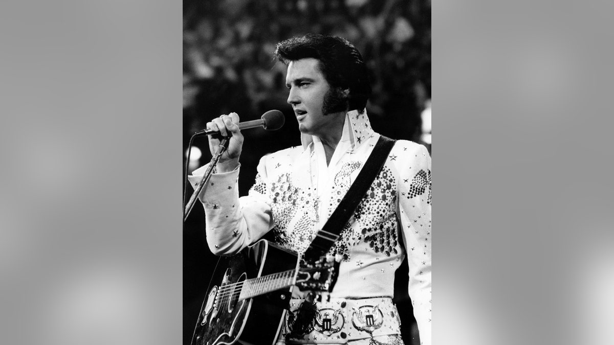 Elvis Presley holding a mic while wearing a white jumpsuit