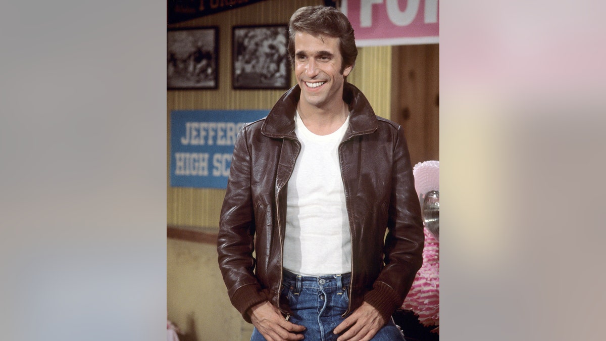 Henry Winkler wearing a brown leather jacket, a white shirt and blue jeans