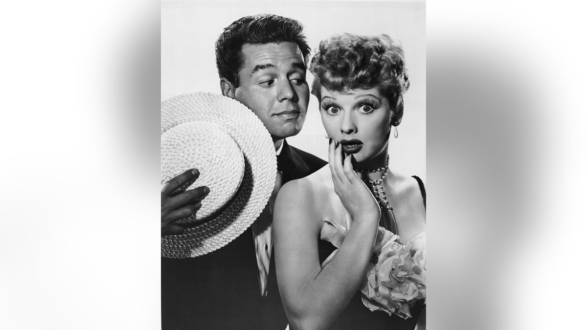 Lucille Ball looking shocked as Desi Arnaz looks at her holding a straw hat