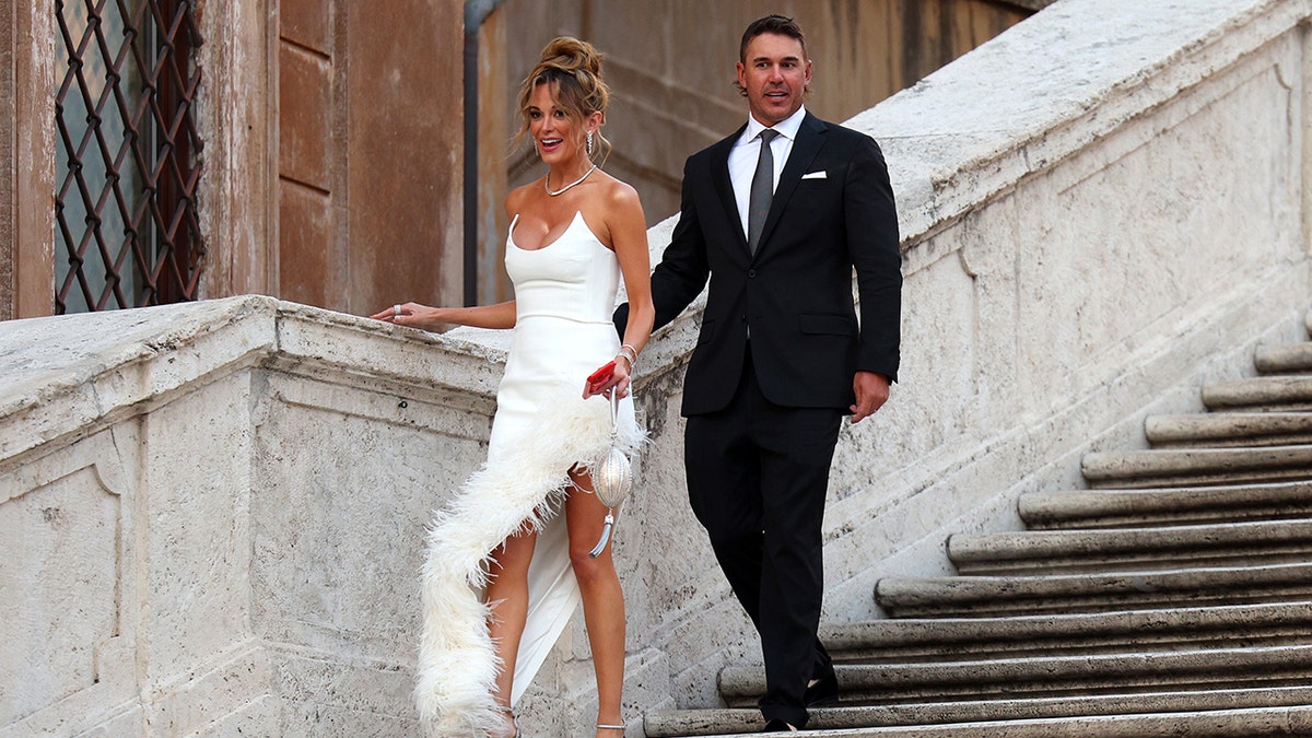 Jena Sims usando um vestido branco e Brooks Koepka vestindo um terno enquanto descem a Escadaria Espanhola