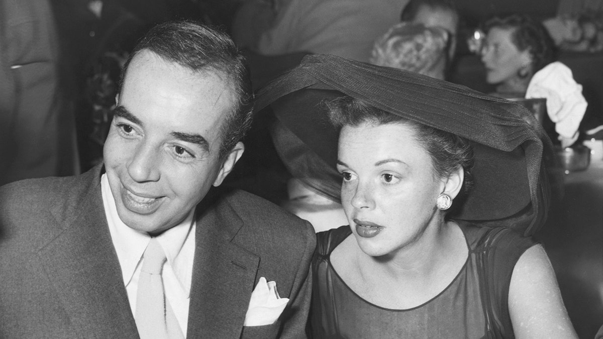 Judy Garland looking serious in a black dress and matching hat next to her husband
