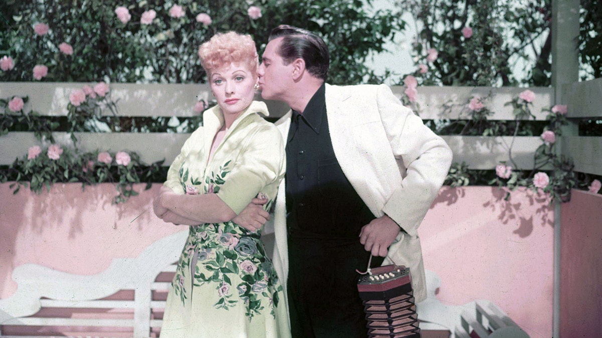 Lucille Ball looking stern as Desi Arnaz kisses their ear as theyre surrounded by pink roses