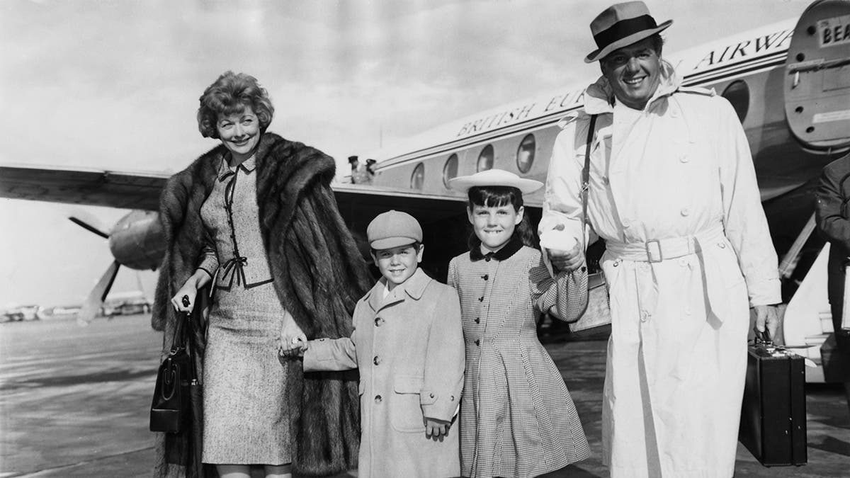 Lucille Ball and Desi Arnaz wearing coats as they hold onto their two young children outside a plane