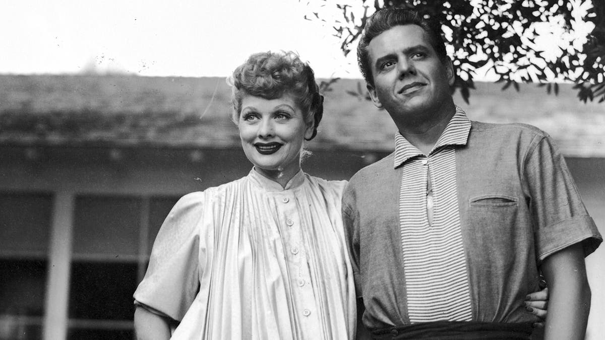Lucille Ball smiling in a white dress while Desi Arnaz looks more serious in front of their home