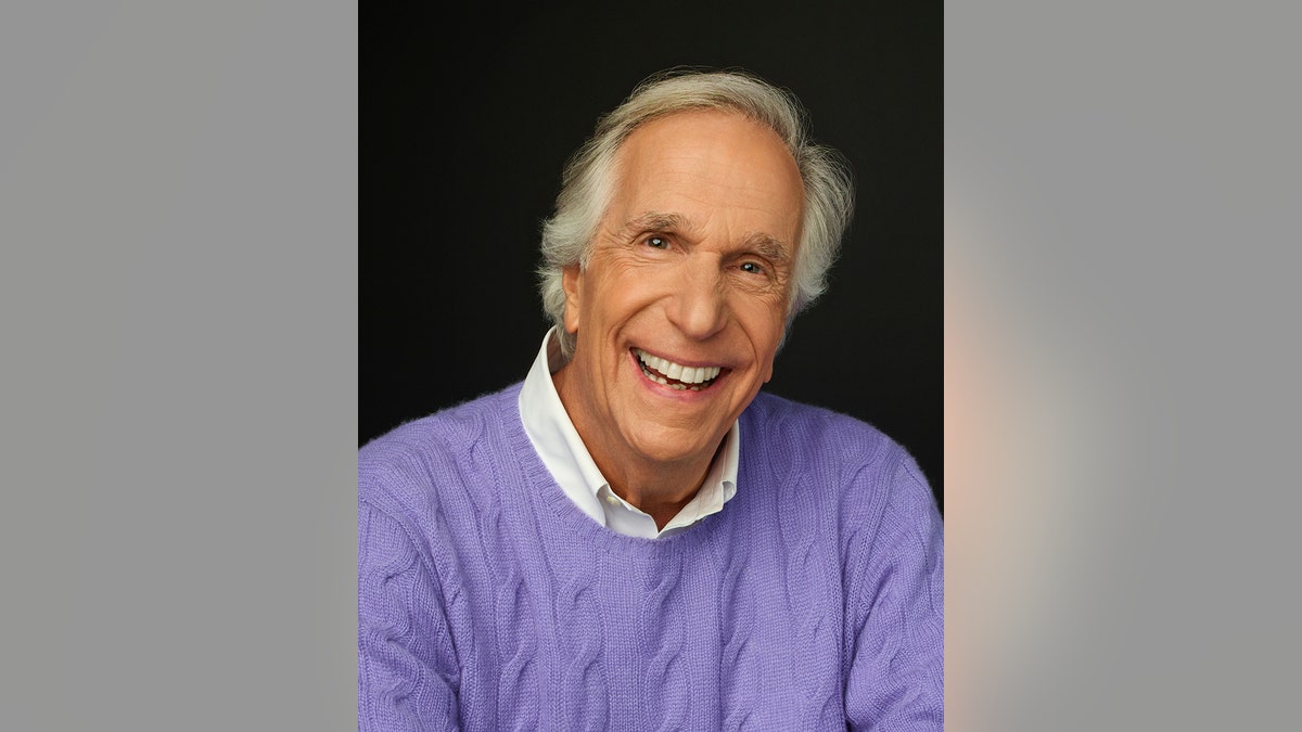 Henry Winkler smiling wearing a purple sweather and a white collared shirt
