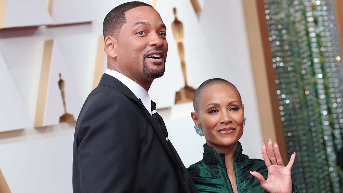 Will Smith olha ligeiramente para cima no tapete do Oscar com Jada Pinkett Smith sorrindo ao lado dele em um vestido verde