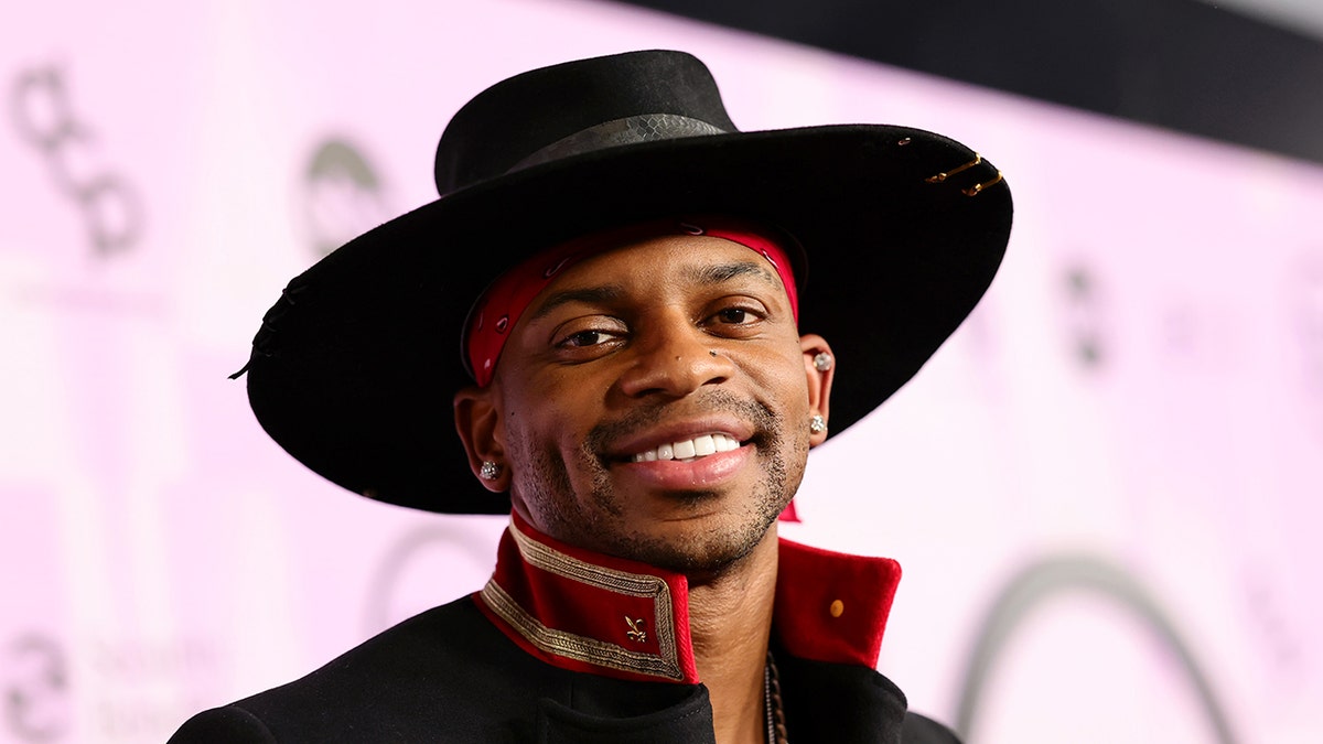 Jimmie Allen smiles at the AMA music Awards in a wide brim black hate and black jacket with a red collar