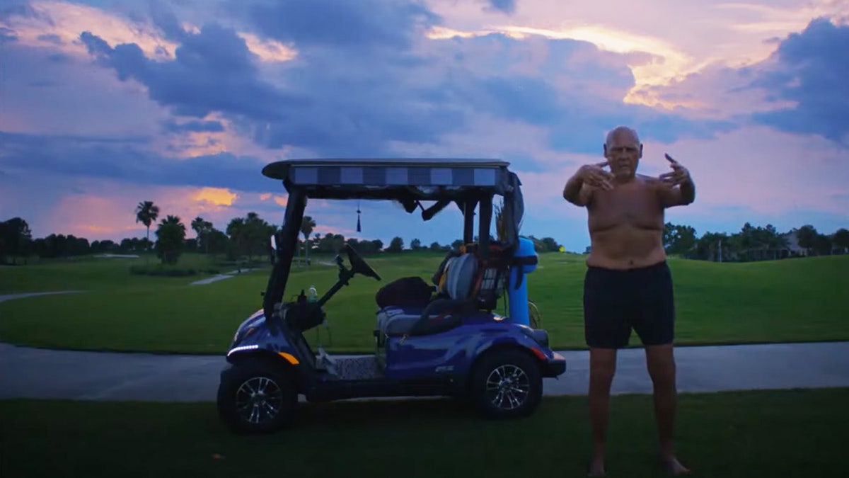 Man stands shirtless next to golf cart performing martial arts exercises.