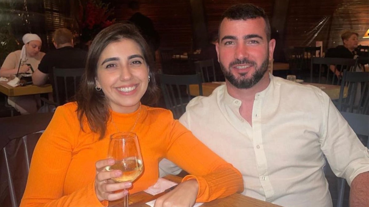 Woman, wearing an orange shirt, drinks a glass of wine as she poses with boyfriend.