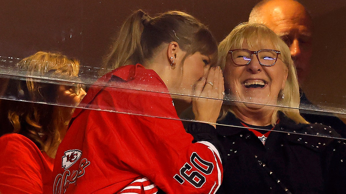 Taylor Swift in a red jacket leans over to whisper something into Donna Kelce's ear who is laughing