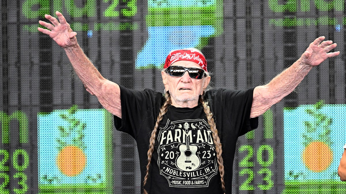 Willie Nelson wearing sunglasses with arms raised on stage