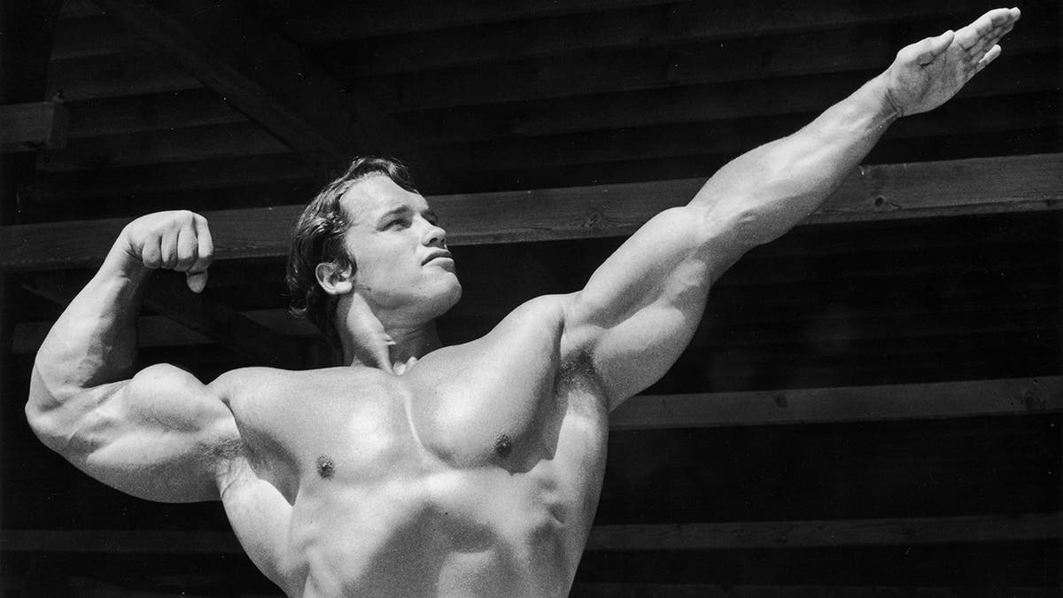 In a black and white photo, Arnold Schwarzenegger flexes one arm and outstretches the other 