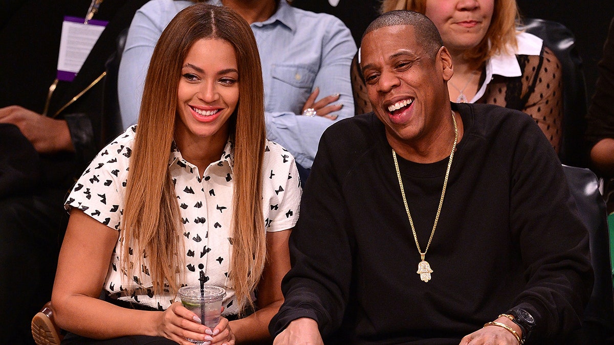 Beyoncé dans une chemise blanche à manches boutonnées est assise au bord du terrain à côté de Jay-Z dans une chemise noire à manches longues