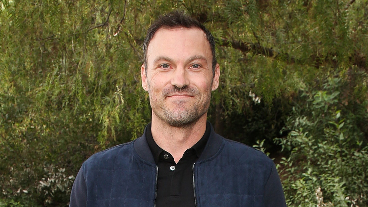Brian Austin Green in a blue jacket over a black shirt soft smiles in front of a tree