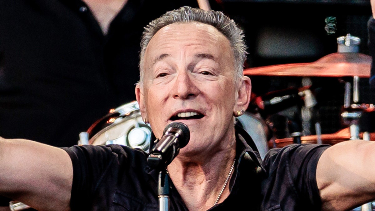 Close up of Bruce Springsteen in a black shirt puts his arms up in the air while performing in Italy