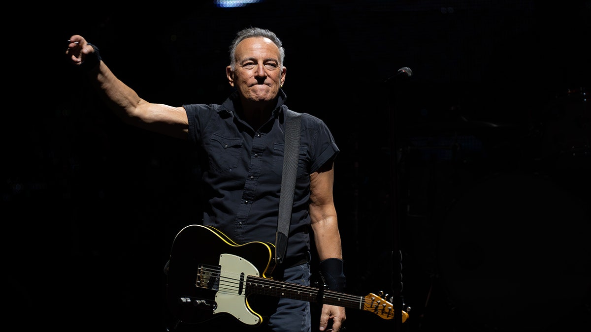 Bruce Springsteen in a black short sleeve button down sticks out his right arm on stage in New Jersey