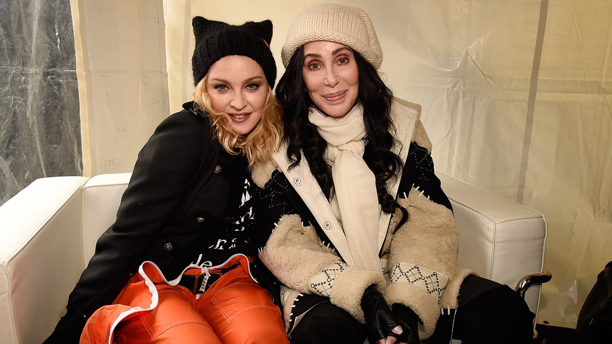Madonna in a black hat and jacket and orange pants smiles for a photo with Cher in a tan hat and jacket